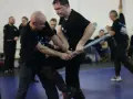 Dwóch mężczyzn w czarnych koszulkach ćwiczy samoobronę z użyciem pałki.