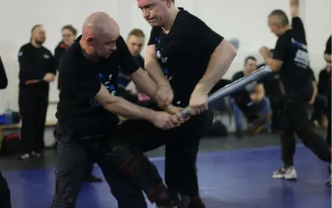 Dwóch mężczyzn w czarnych koszulkach ćwiczy samoobronę z użyciem pałki.