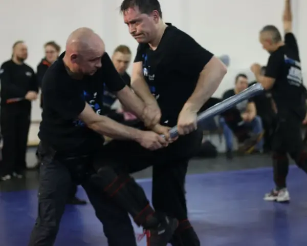 Dwóch mężczyzn w czarnych koszulkach ćwiczy samoobronę z użyciem pałki.